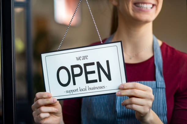 Small business owner smiling while turning the sign for the reopening of the place after the quarantine due to covid-19. Happy businesswoman standing at her restaurant or coffee shop gate with open signboard. Close up of woman"u2019s hands holding sign now we are open support local business.